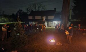 A group of people gathered around a bonfire and a small evergreen tree.
